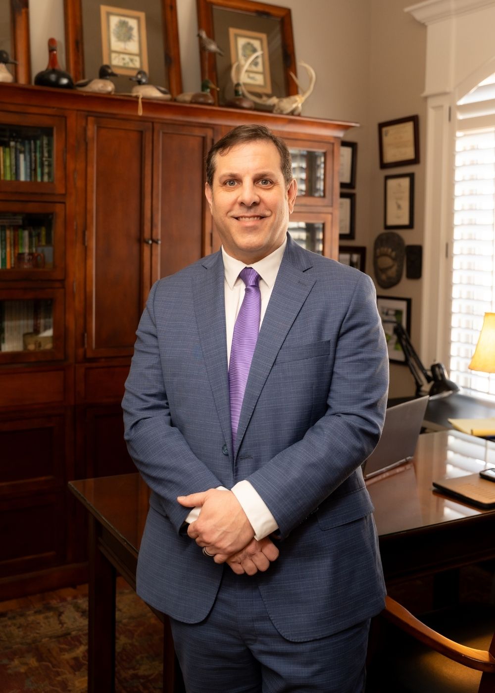 Lunde Office (10) A man in a blue suit and purple tie stands smiling in a wood-paneled office with bookshelves, framed certificates, a desk, and a lamp by the window.