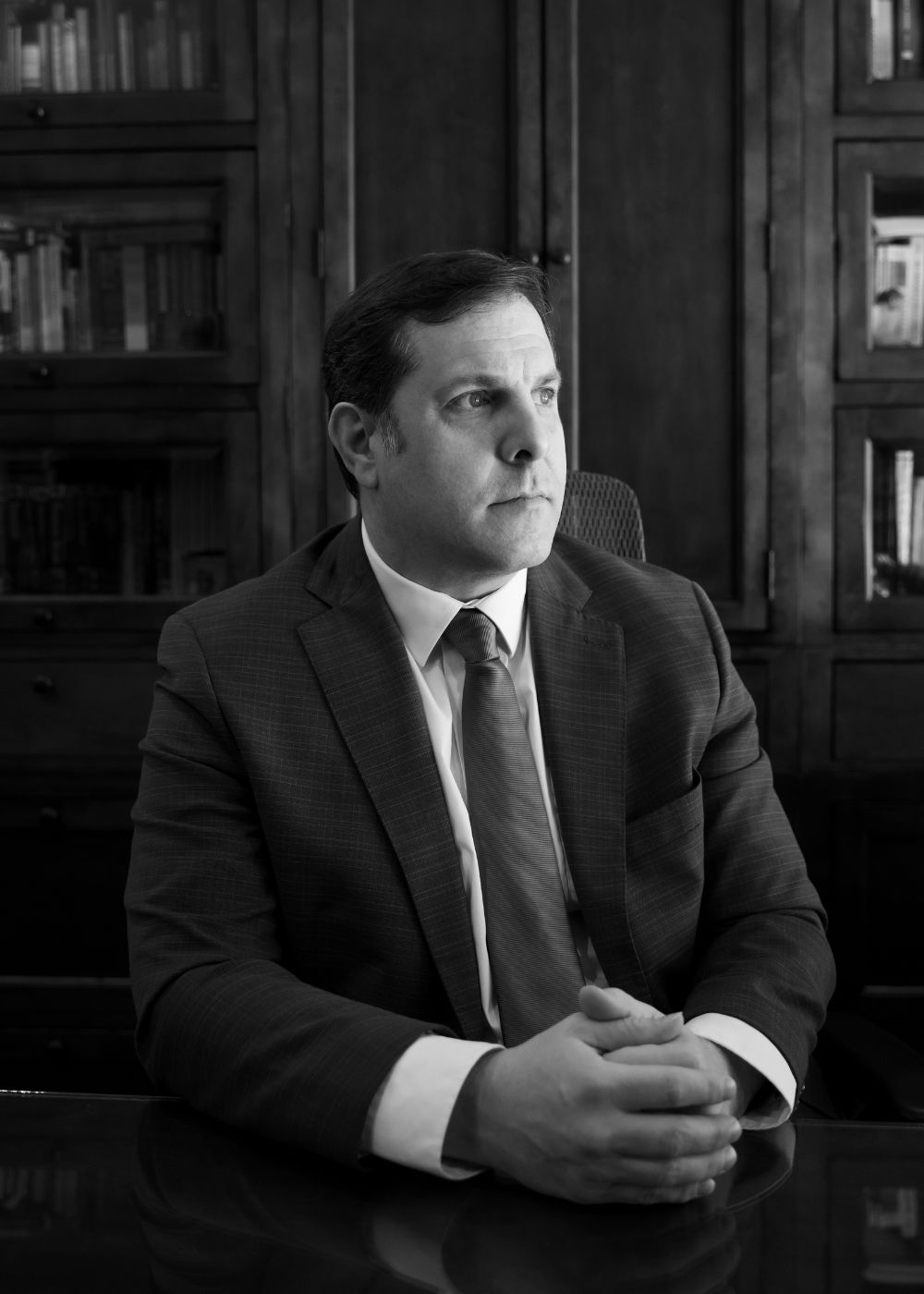 Lunde Office (8) A man in a suit sits at a table with his hands clasped, looking to the side. He is in an office with wooden bookshelves filled with books behind him. The image is in black and white.