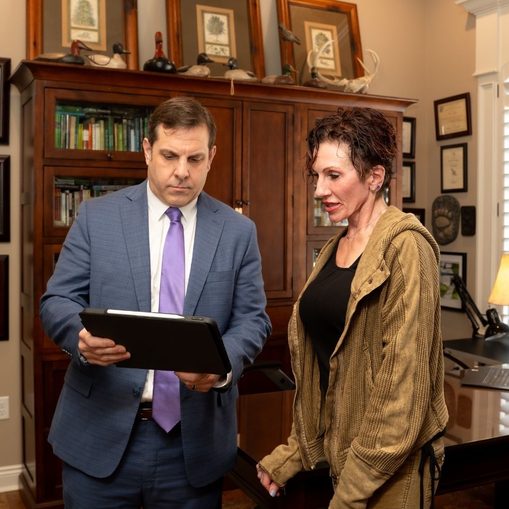 Lunde Services (4) A man in a blue suit shows a document or folder to a woman in casual clothes in a well-furnished office with bookshelves, framed certificates, and decor in the background.