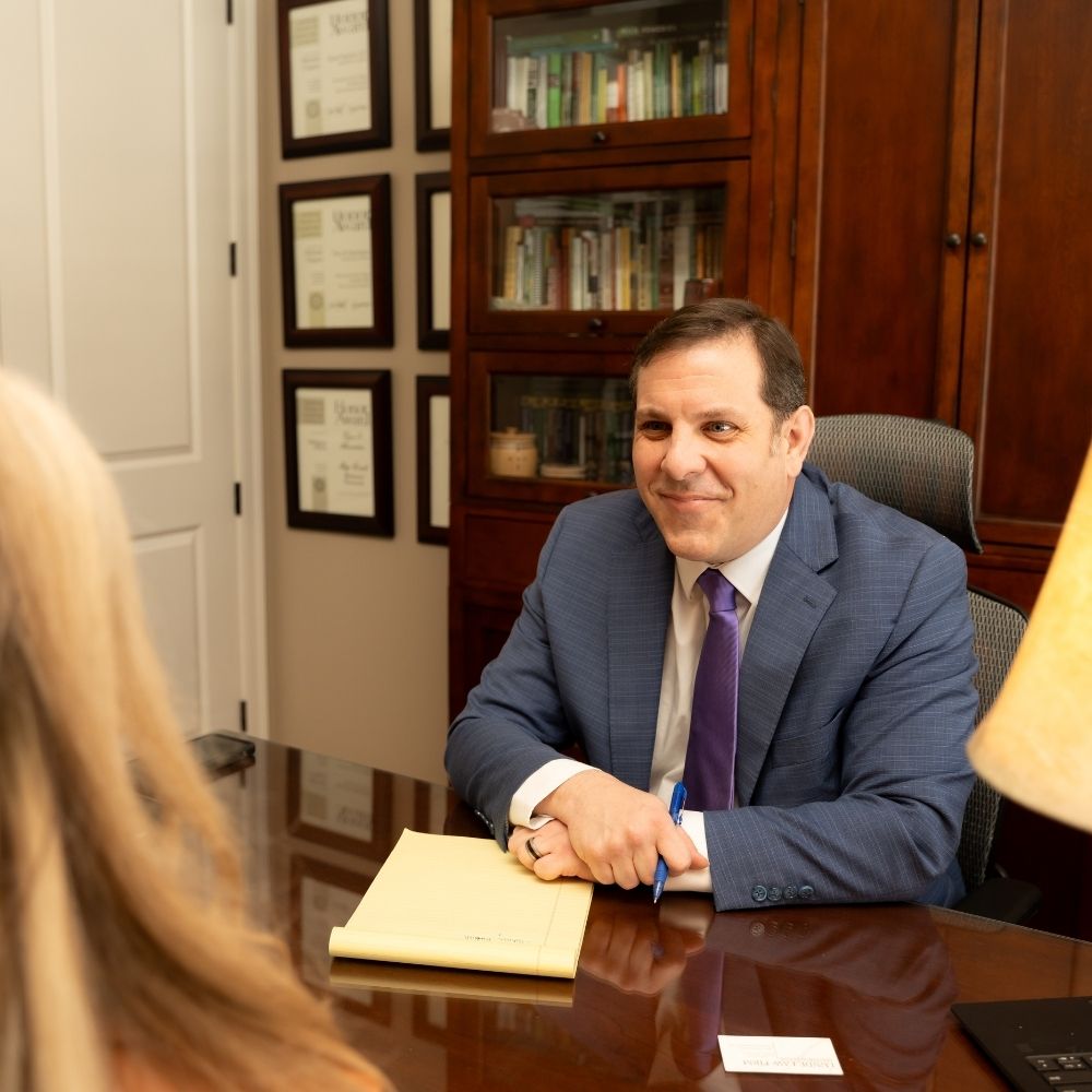 A man in a blue suit sits at a desk, smiling and talking to a woman. He holds a pen and has a yellow notepad in front of him. Behind him are bookshelves and framed certificates on the wall.