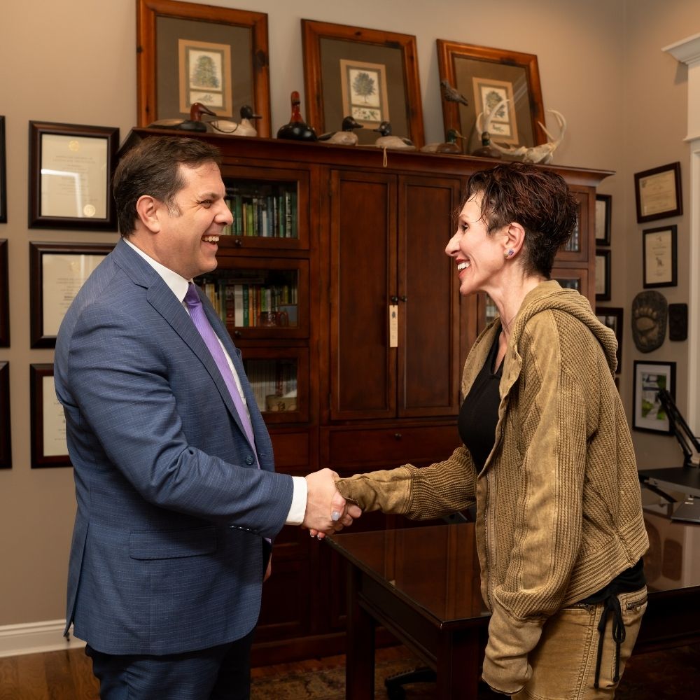 Lunde Thank you (3) A man in a blue suit and a woman in a brown jacket shake hands and smile at each other in an office with wood furniture, framed certificates, and artwork on the walls.
